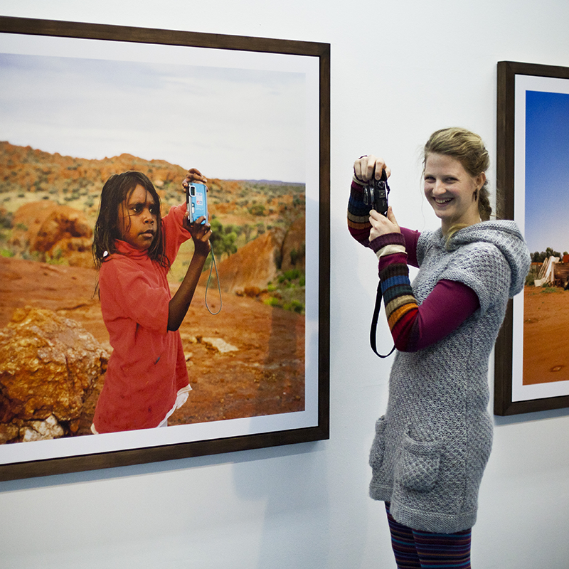 Die Fotografin Mira Unkelbach steht vor einem Bild ihrer Diplomarbeit, lacht und hält eine Kamera in der Hand. Auf dem Bild an der Wand ist ein Walpiri Mädchen abgebildet, welche eine blaue Kamera in den Händen hält.