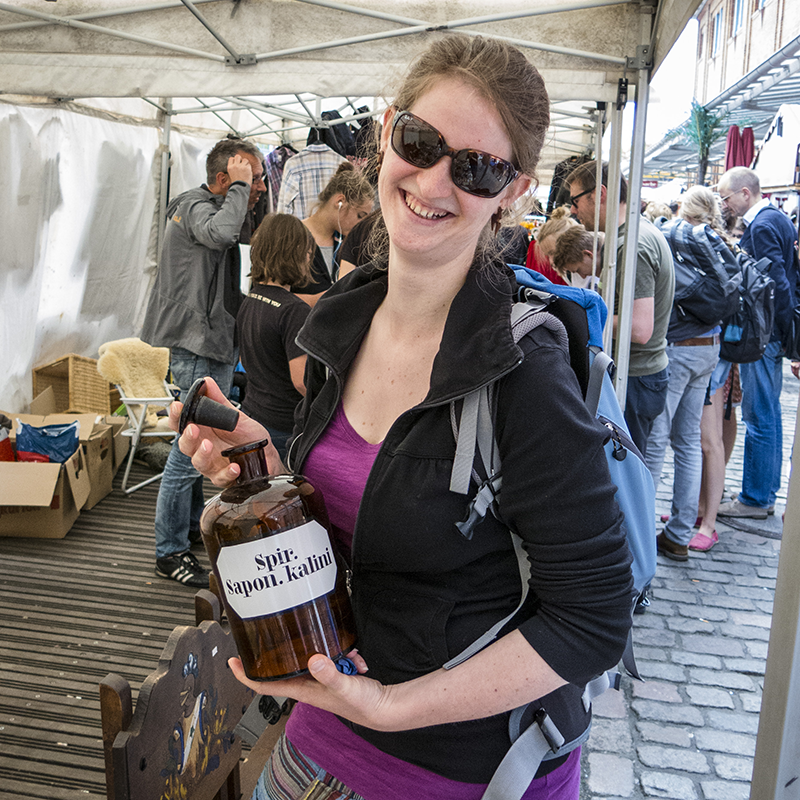 Die Fotografin Mira Unkelbach steht auf einem Flohmarkt und hat ein altes Apothekerglas in ihrer Hand. Sie trägt eine Sonnenbrille und einen Rucksack auf dem Rücken. Außerdem grinst sie glücklich in die Kamera.