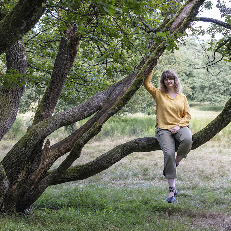Die Fotografin Mira Unkelbach sitzt auf dem Ast eines alten und niedrigen Baums. Sie trägt eine olive-farbende Hose und eine gelbe Bluse und guckt in die Kamera.