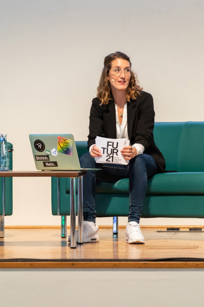 Eventfotografie. Eine junge Frau sitzt auf einem grünen Sofa. Vor ihr steht ein Tisch mit einem aufgeklappten Laptop. In der Hand hält sie ihre Moderationskarten für die Veranstaltung.