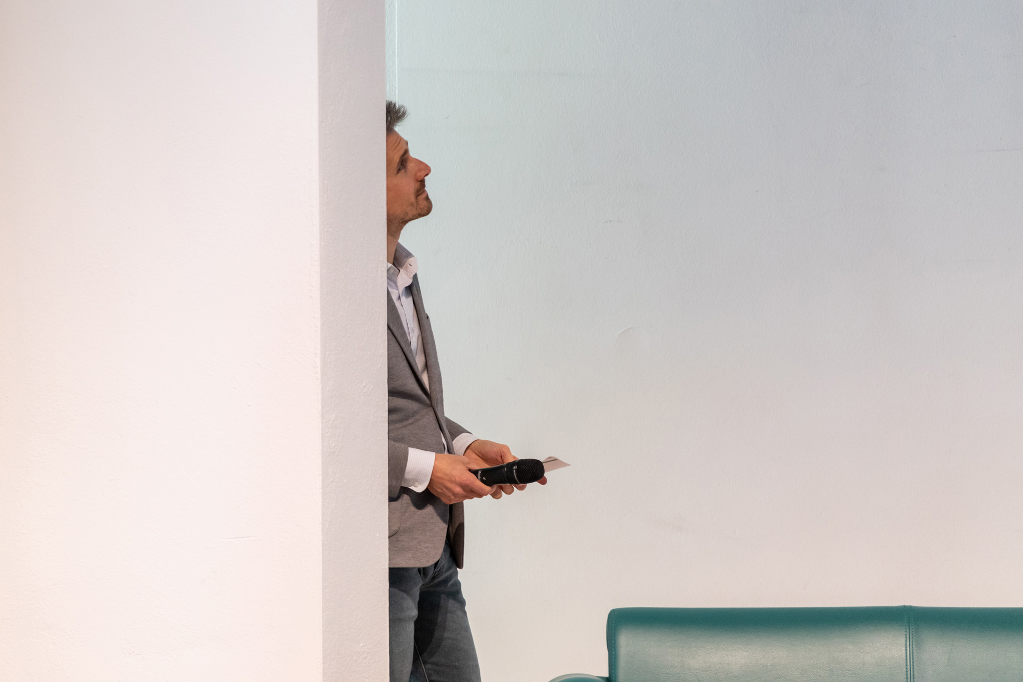 Veranstaltungsfotografie. Ein Mann steht an eine Wand gelehnt und guckt leicht nach oben. In seinen Händen hält er ein Mikrofon und Moderationskarten. Vor ihm steht ein grünes Sofa, welches nur im Anschnitt erkennbar ist.