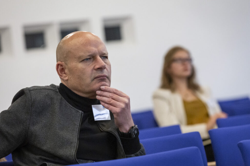 Ein Mann mittleren Alters bei einer Tagung. Er sitzt auf einem blauen Stuhl. Im Hintergrund sieht man eine junge Frau, die ebenfalls im Publikum sitzt. Der Mann hat seine Hand am Kinn und macht einen nachdenklichen Eindruck.