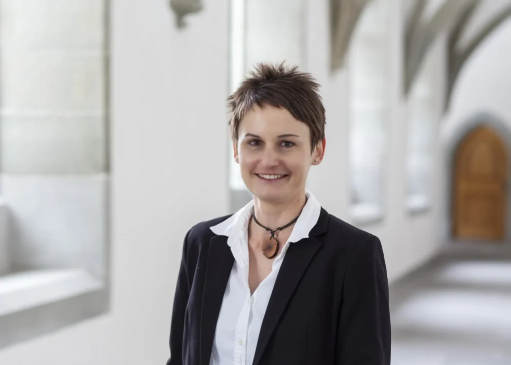 Portraitfotografie in Bonn - Eine Frau steht im Flur eines Altbaus. Sie trägt eine weiße Bluse und einen schwarzen Blazer. Im Hintergrund erkennt man eine Holztür.